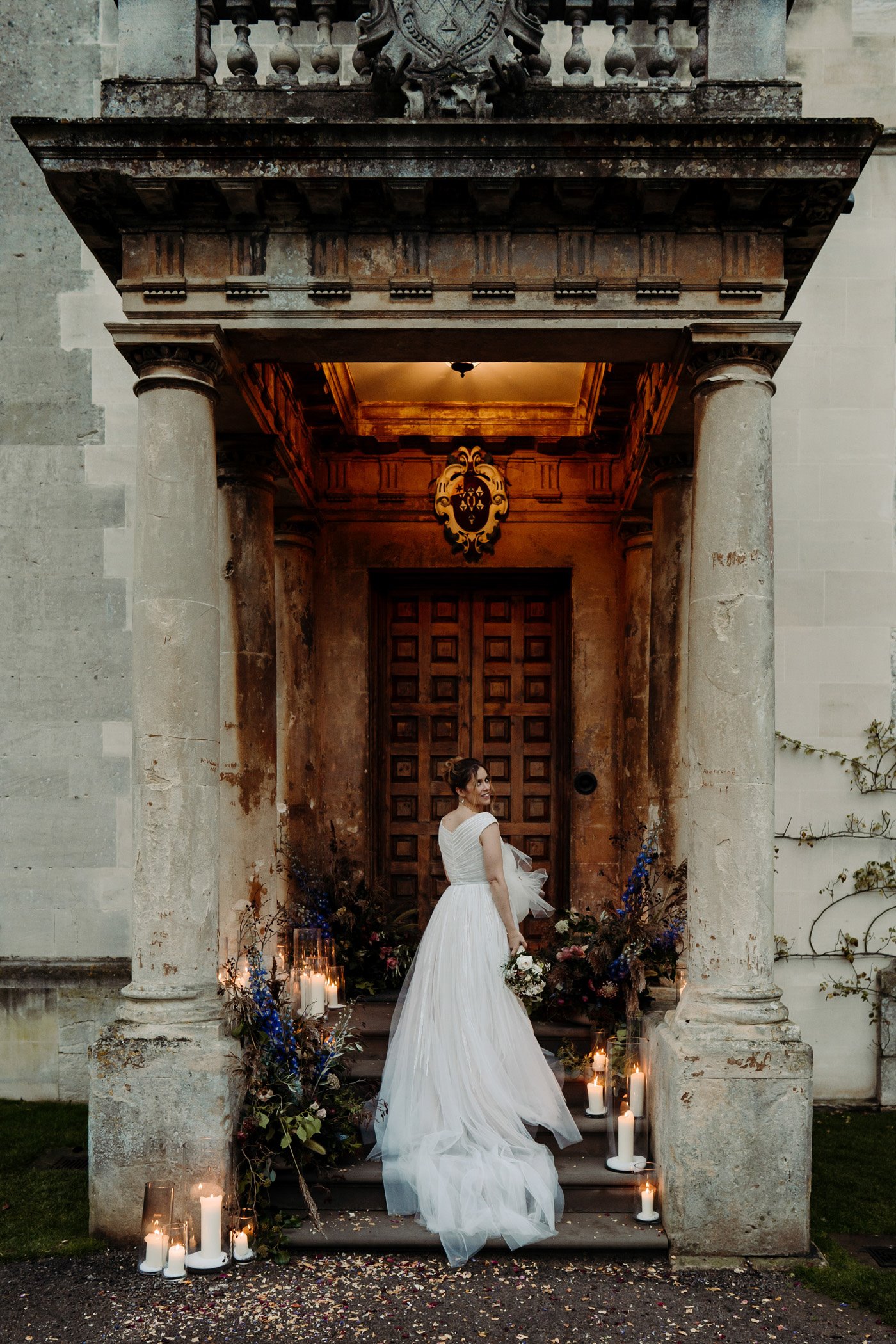 Bride stands on the entrance portico of Elmore Court with her long white gown trailing down the steps. Candles and flowers decorate either side of her