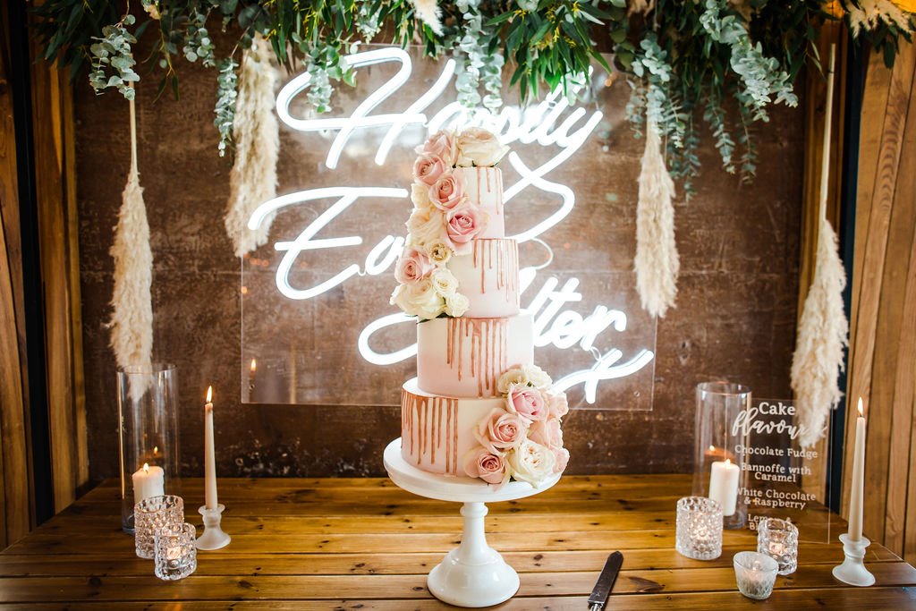 Four tier asymmetric pink wedding cake decorated with real flowers, beautifully presented on a cake table with neon sign saying happily ever after and greenery and pampas grass hanging above