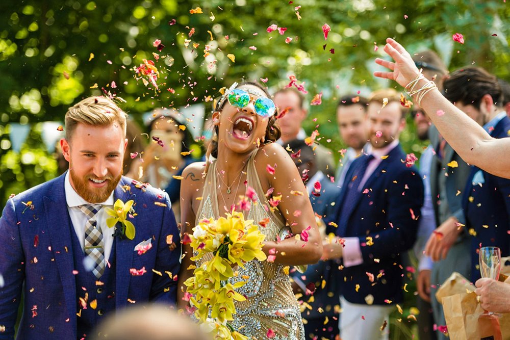 Festival bride wearing sunglasses and holding a yellow bouquet throws back her head and laughs in an explosion of confetti as her newlywed husband keeps his head down