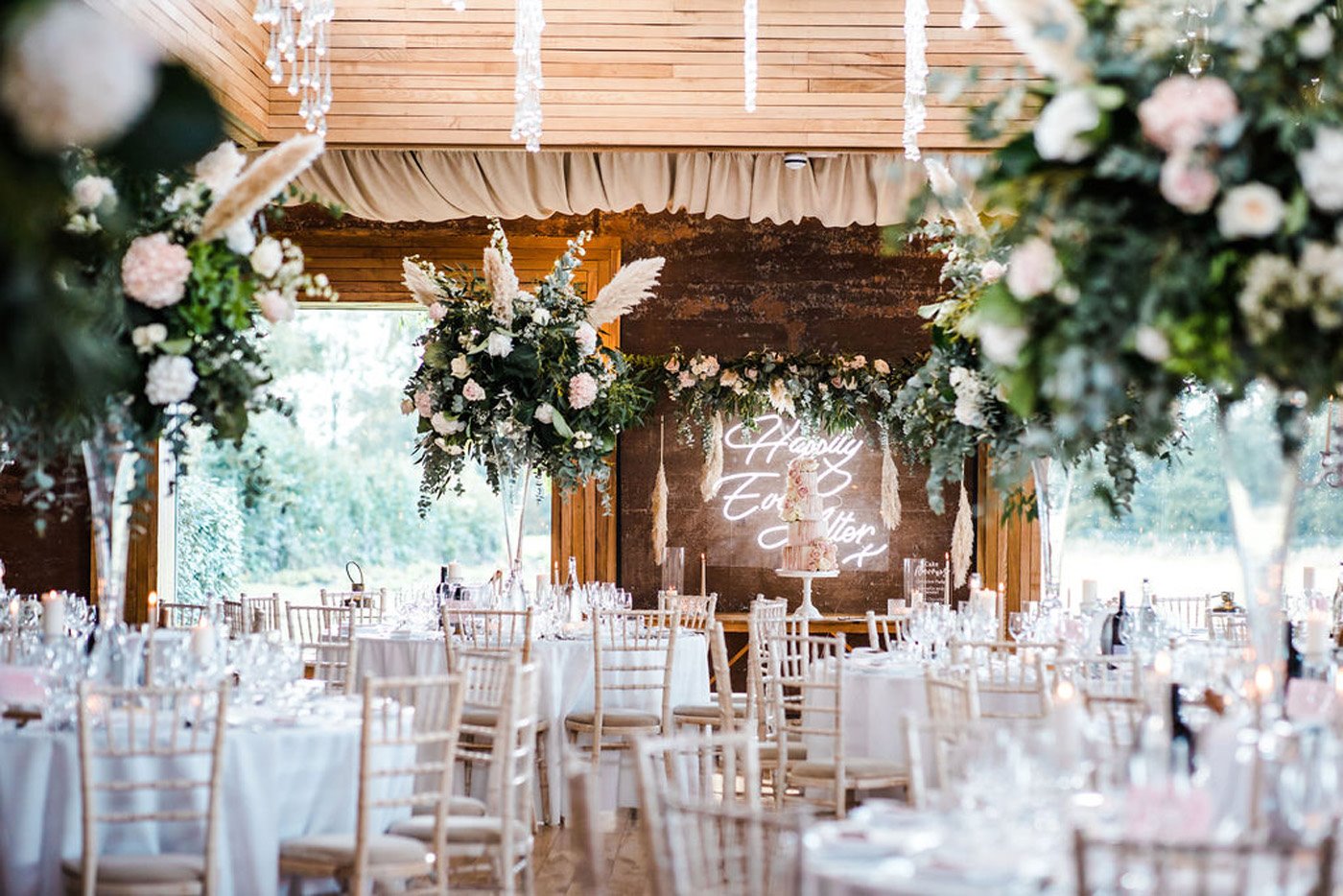 Pretty pink and greenery wedding reception with round tables and foliage pampas grass floral arrangements, neon sign back lighting the cake table