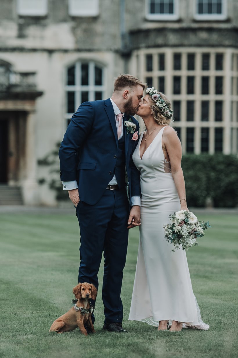 Dog friendly micro wedding at Elmore court. Bride and Groom kiss on the lawn in front of stately home with their puppy bridesmaid dressed in flowers beside them