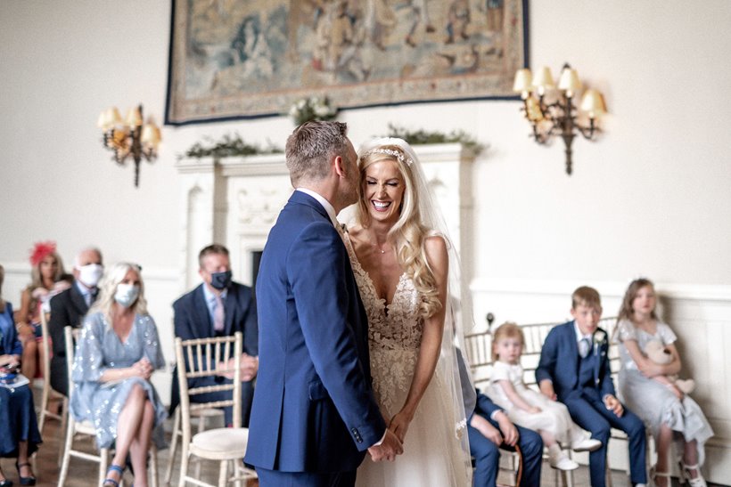 Mature bride in pearls tiara and lace wedding gown and her groom stand at end of aisle holding hands smiling in stately home hall of Elmore Court