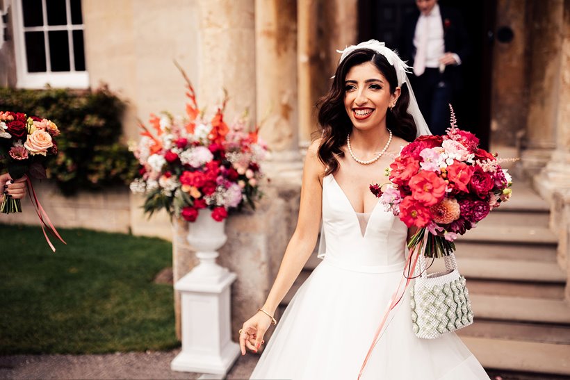 Beautiful modern bride in pearl accessories; headpiece and pearl necklace holding bright pink and red bouquet and pearl handbag outside her glamorous wedding venue 