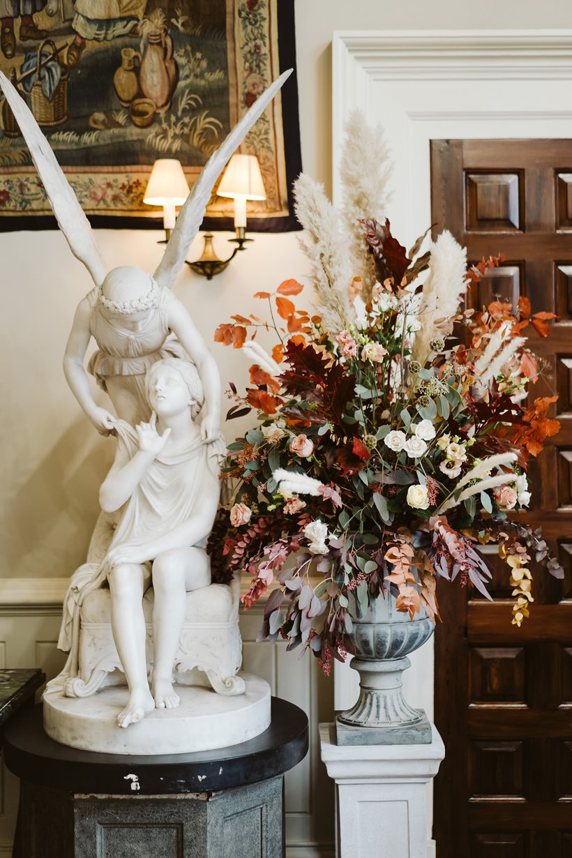 Autumn foliage, flowers and pampas grass in a large urn next to angel statues at elmore court in gloucestershire