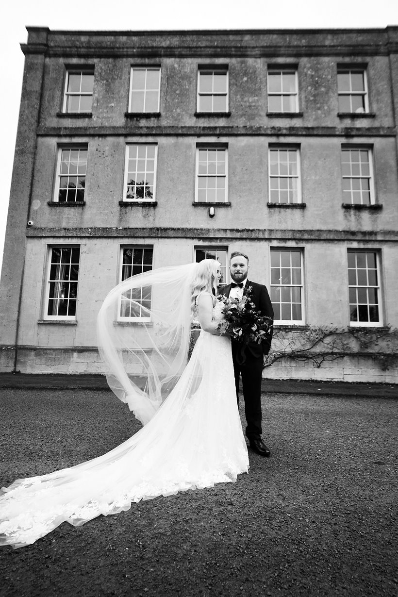 Moody black and white photo of Bride and groom standing in front of spooky old mansion house on their Halloween wedding in England