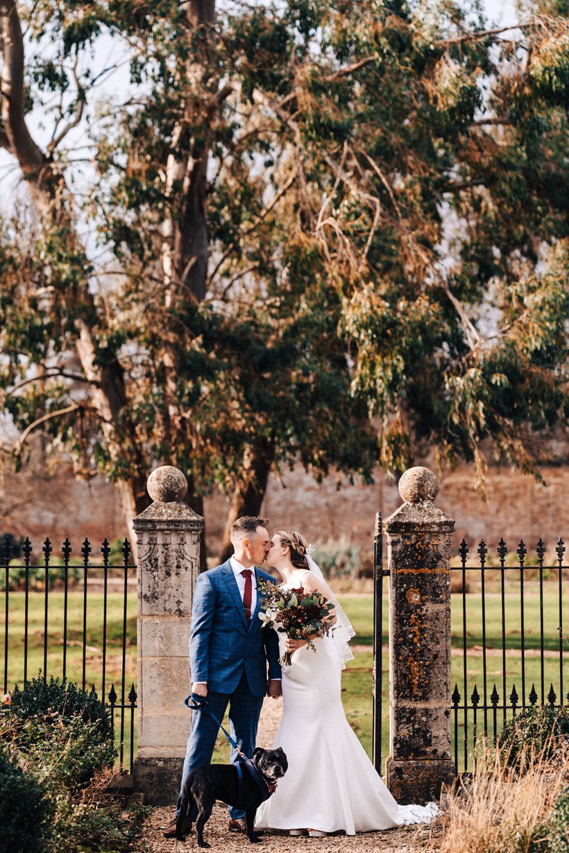 bride and groom kiss with their dog on a lead in the walled garden at their christmas wedding at elmore court