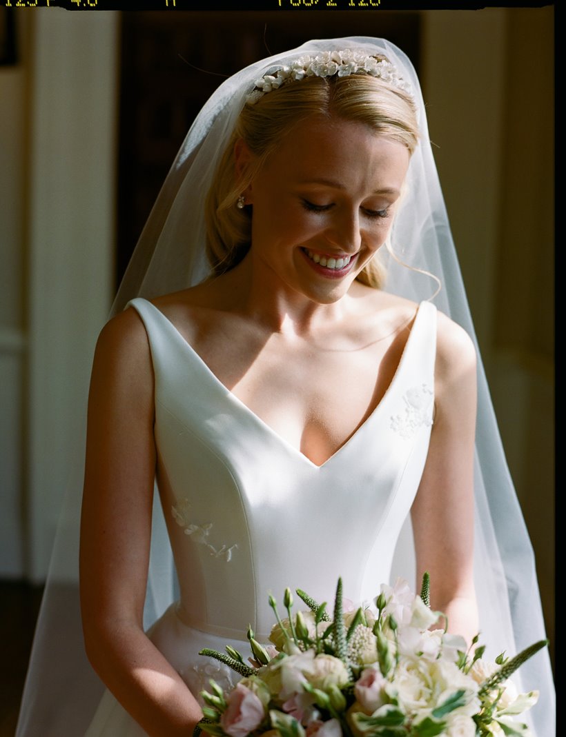 Demure and elegant bride in chic simple satin wedding gown, pearl tiara and veil, pearl necklace and pearl earrings smiles down at her bouquet of white, pale pink and green flowers