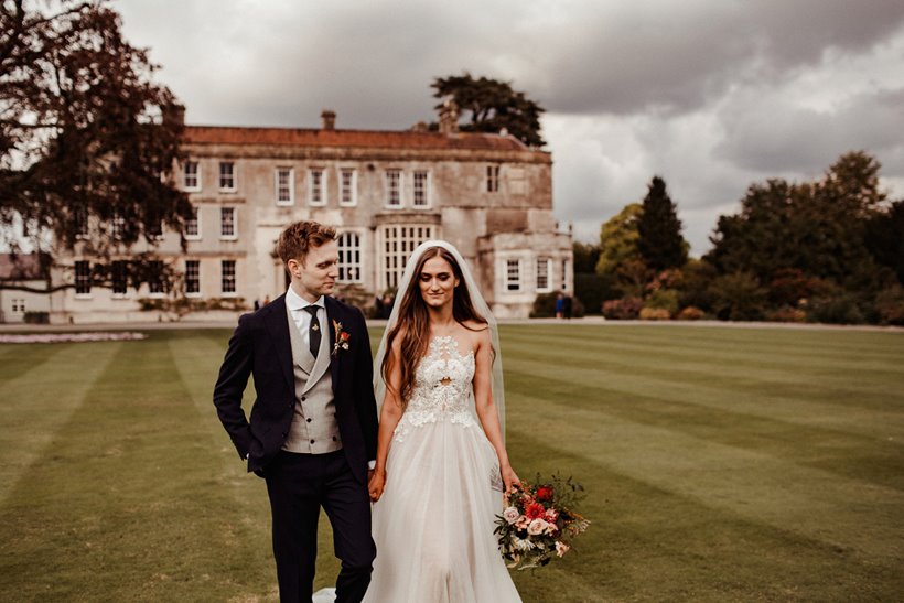 Pre raphaelite inspired bride stands outside mansion house wedding venue with her groom on the lawn