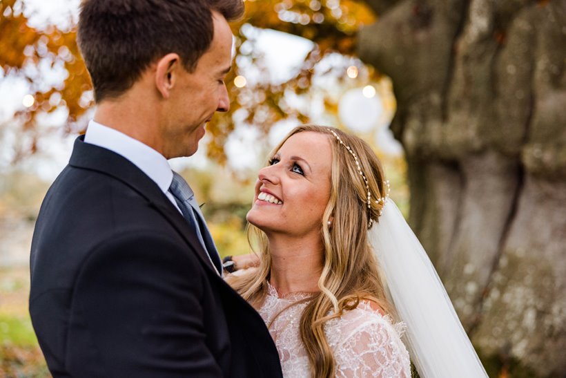 Bride in pearl headpiece and veil and groom stand underneath copper beech with golden leaves look lovingly at eachother 