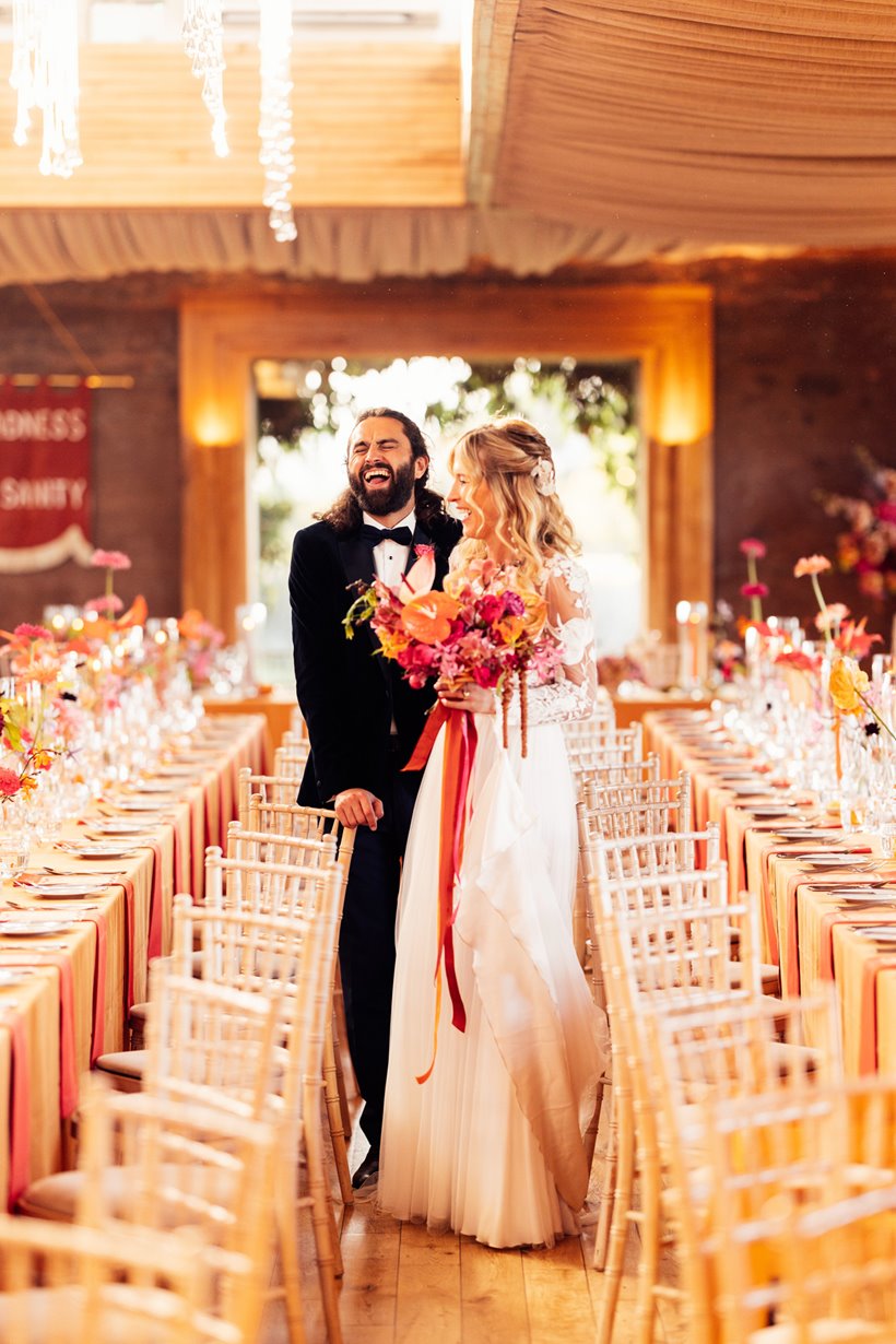 Fun loving boho bride and groom laugh with joy at their colourful wedding reception with bright flowers in the cotswolds