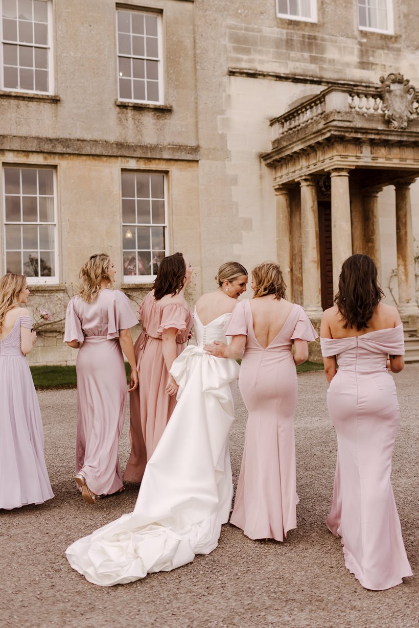 Bride in dress with huge bow and bridesmaids in pink dresses figure hugging with backs to the camera in front of stately home wedding venue