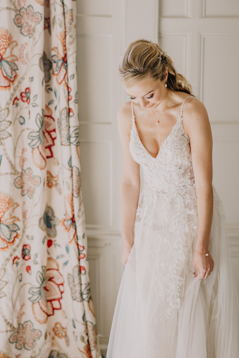 Blonde bride in pearl earrings, necklace and simple pearl bracelet looks down at her lace dress against embroidered curtains and wood panelling of stately home wedding venue