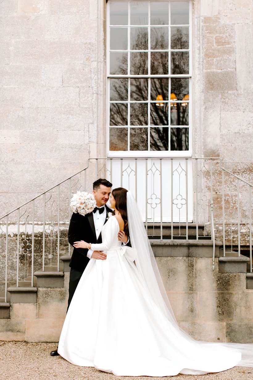 Bride and groom embrace next to steps of a stately home in Gloucestershire